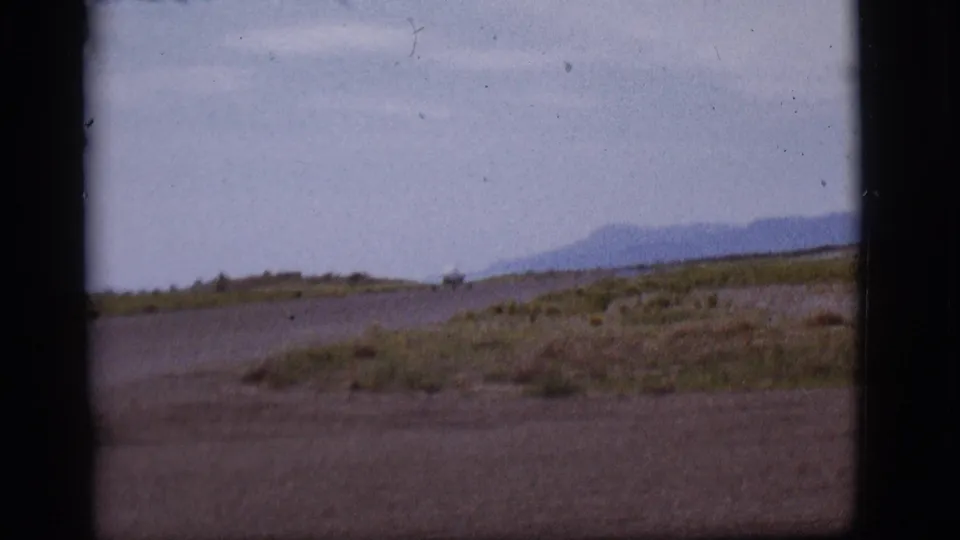 A wide 8mm view of the Flagstaff runway area shows the small plane farther away against dry grass and a mountain backdrop.