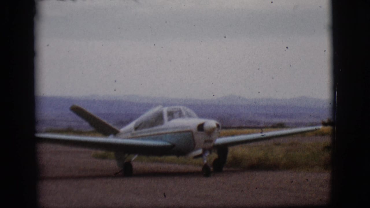 Small white and blue airplane taxiing beside a grassy runway with hills in the distance.