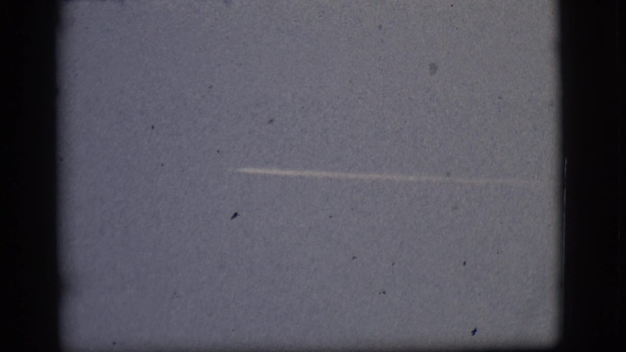 Distant jet contrail crossing a pale blue sky with dark film-frame borders.
