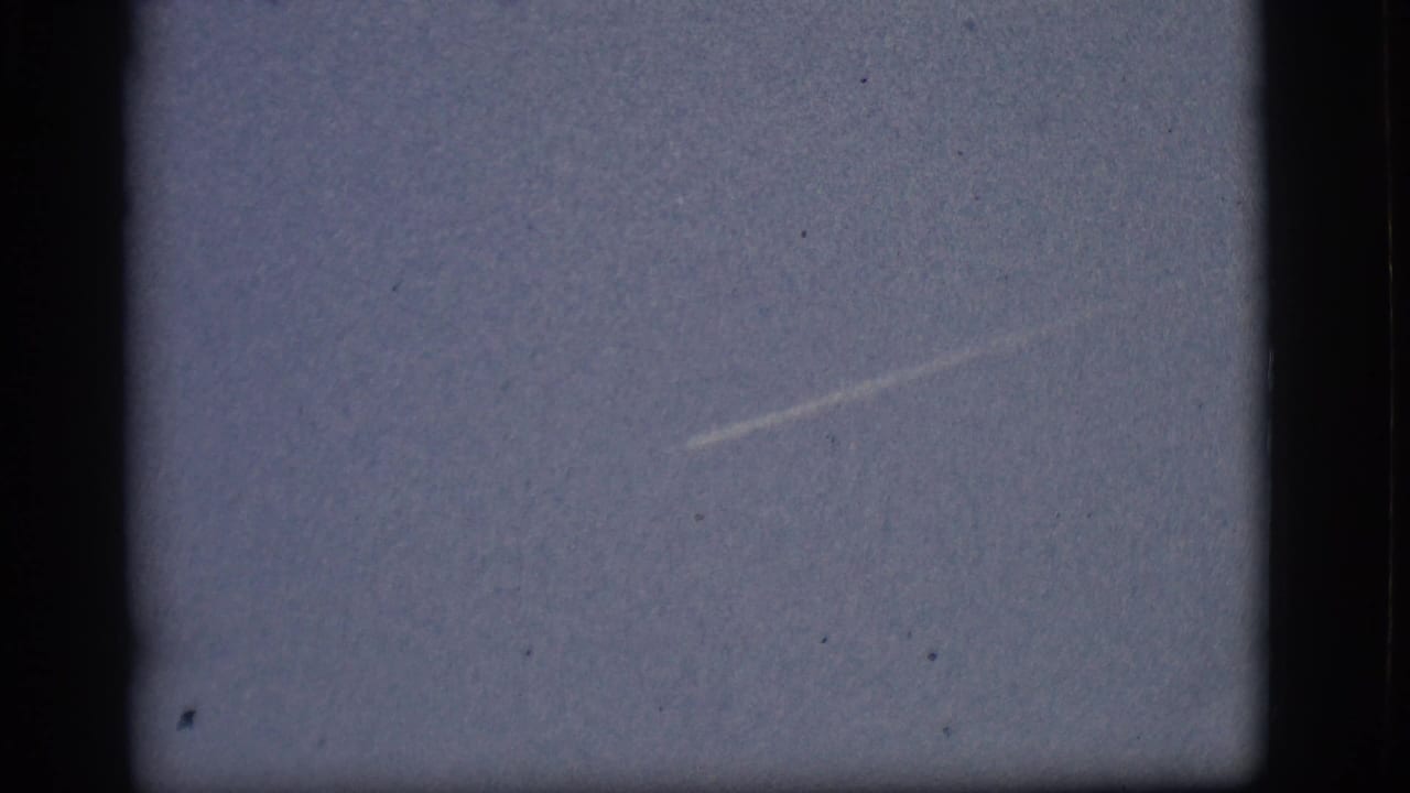 Distant aircraft or bright point leaves a thin contrail across pale blue sky.