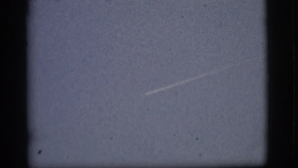 1968 Flagstaff home-movie frame showing a small distant aircraft or bright point with a faint diagonal contrail in pale sky.