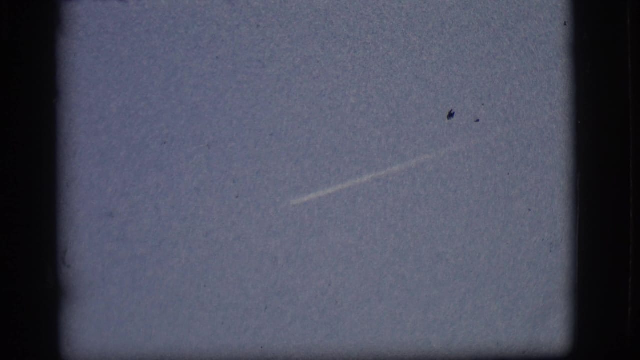 1968 Flagstaff sky frame with a faint diagonal contrail and small distant aircraft trace, seen through dark 8mm gate edges.