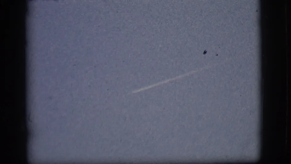 1968 Flagstaff sky frame with a faint diagonal contrail and small distant aircraft trace, seen through dark 8mm gate edges.