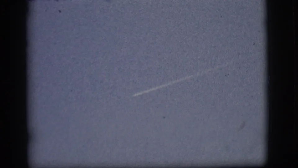 1968 Flagstaff home-movie still with a fading diagonal aircraft contrail across a soft, grainy blue sky.