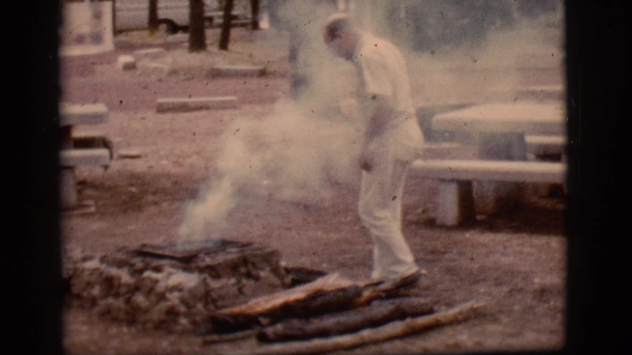 Man in white clothing stands near a smoky campfire at a campground.