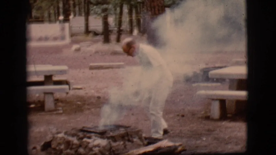 A man in light clothing bends toward a smoky fire pit in the 1968 Flagstaff campground scene, with picnic tables and pine trees visible nearby.