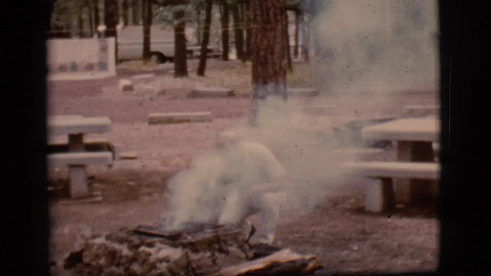Smoke drifts across the 1968 Flagstaff campground frame as the man bends low over the fire pit near logs, rocks, picnic tables, and trees.