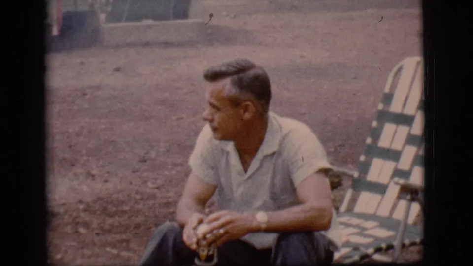 1968 Flagstaff, Arizona home-movie frame showing a seated man holding a drink near a striped folding chair on bare campground ground.