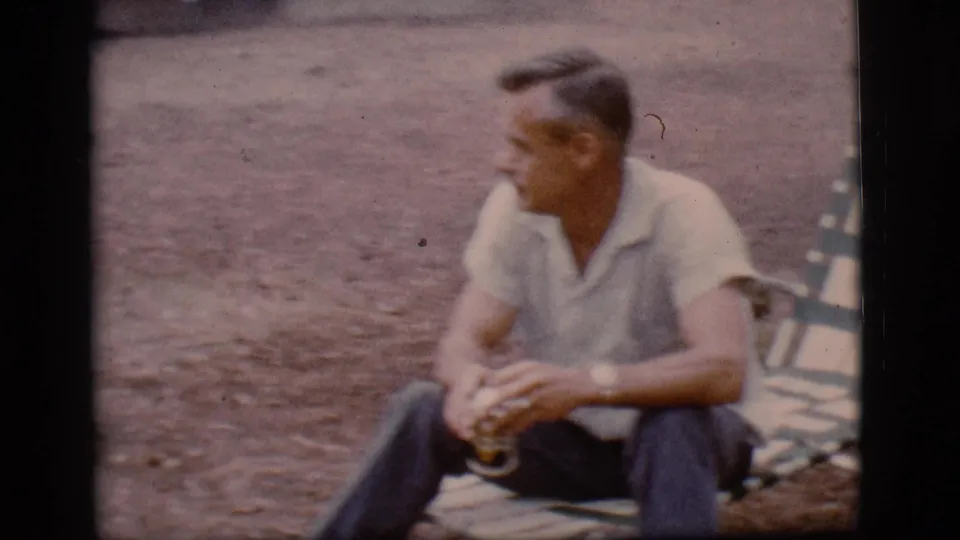 1968 Flagstaff, Arizona frame with a man seated at a campsite, hands around a drink, with a striped folding chair beside him.