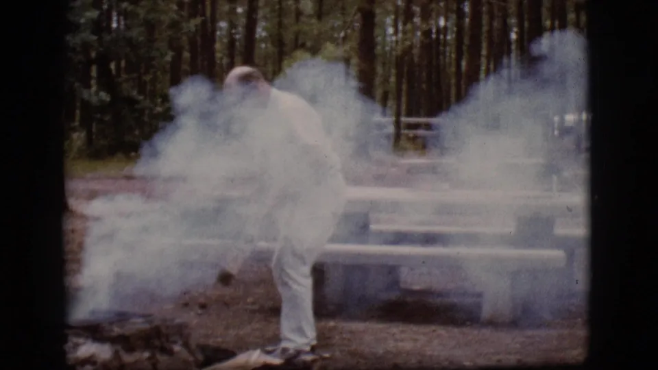 The man bends into dense campfire smoke at a wooded Flagstaff campsite, with picnic tables visible behind him.
