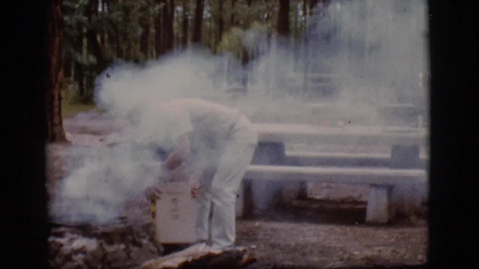 At the wooded 1968 Flagstaff campsite, the man bends over a container while smoke drifts across the picnic-table area.