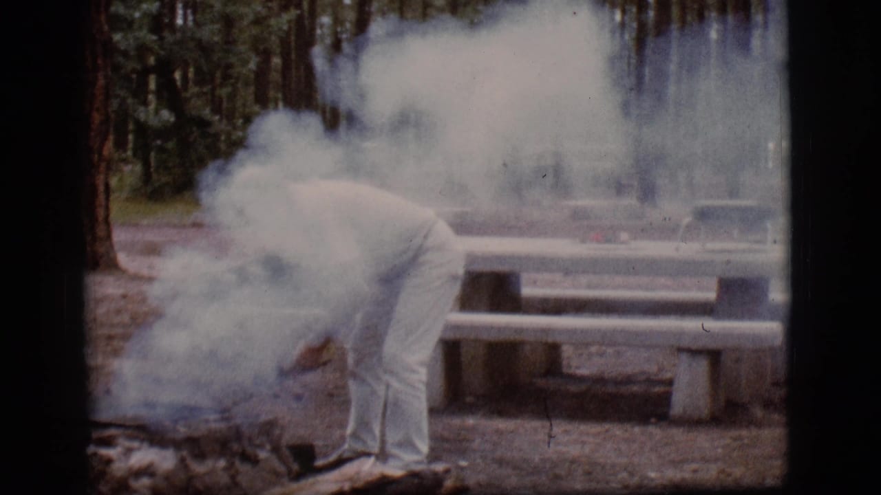 Man in white clothing bends over a smoky campfire ring near a concrete picnic table.