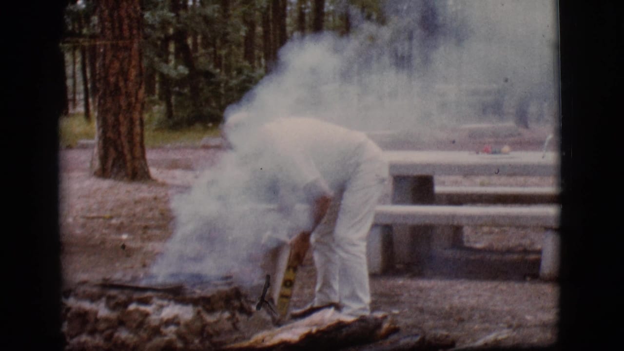 The man remains bent over the smoldering fire pit as white smoke drifts across the Flagstaff campground scene.