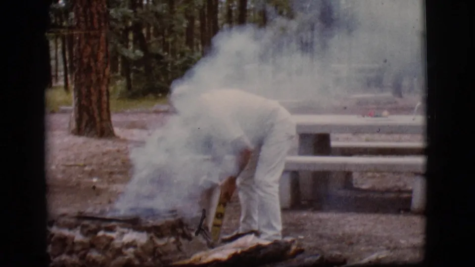 The man remains bent over the smoldering fire pit as white smoke drifts across the Flagstaff campground scene.