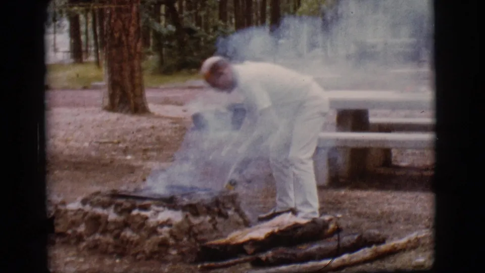 A man in light clothing tends the smoldering campground fire while smoke rises between him, the logs, and a concrete picnic table.