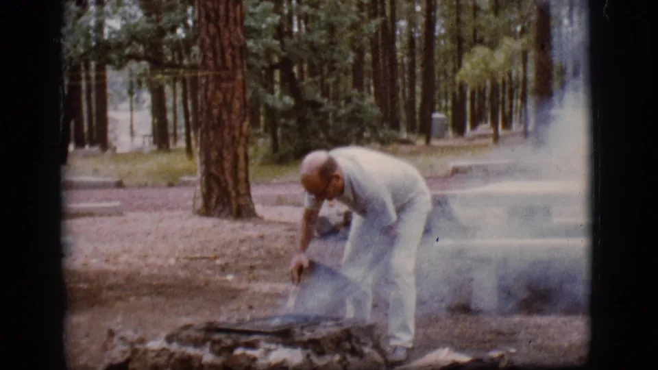 At the end of the clip, the man bends over the low fire pit as smoke trails sideways through the 1968 Flagstaff campground view.