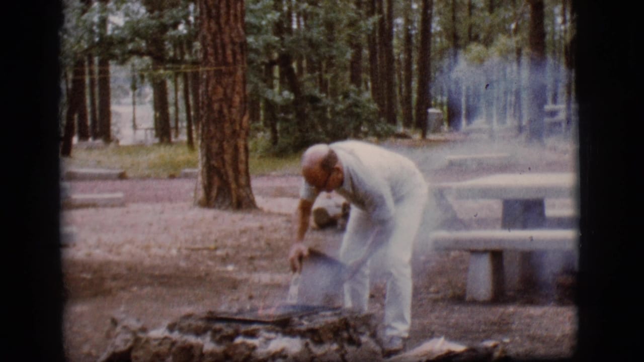 Man bends over a smoky campfire pit with a bucket in a pine campground.