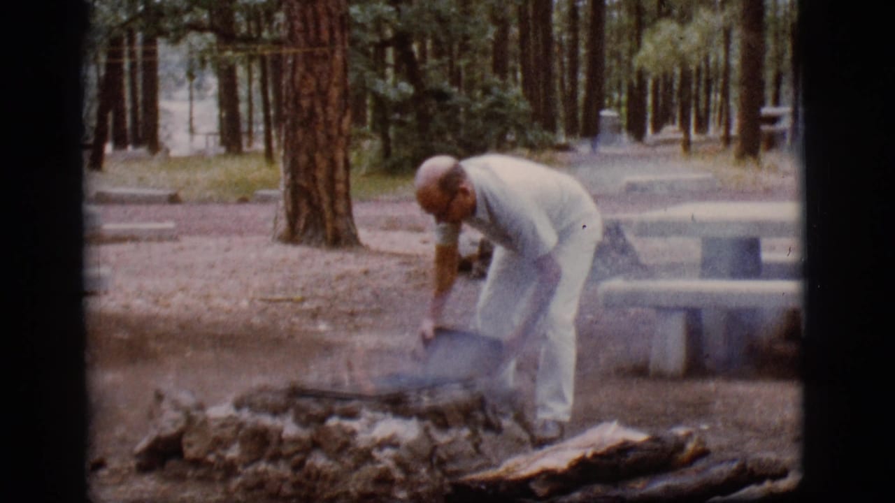 A man in light clothing bends over a smoking campfire pit with a bucket at a wooded Flagstaff campsite in 1968 home-movie footage.