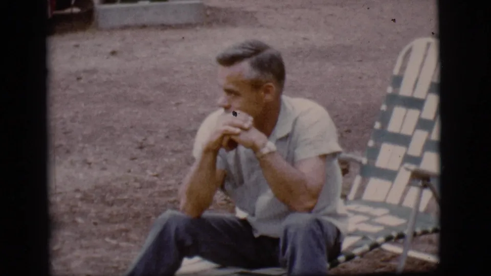 The seated man looks across the campground from a striped folding lounge chair in 1968 Flagstaff, Arizona footage.