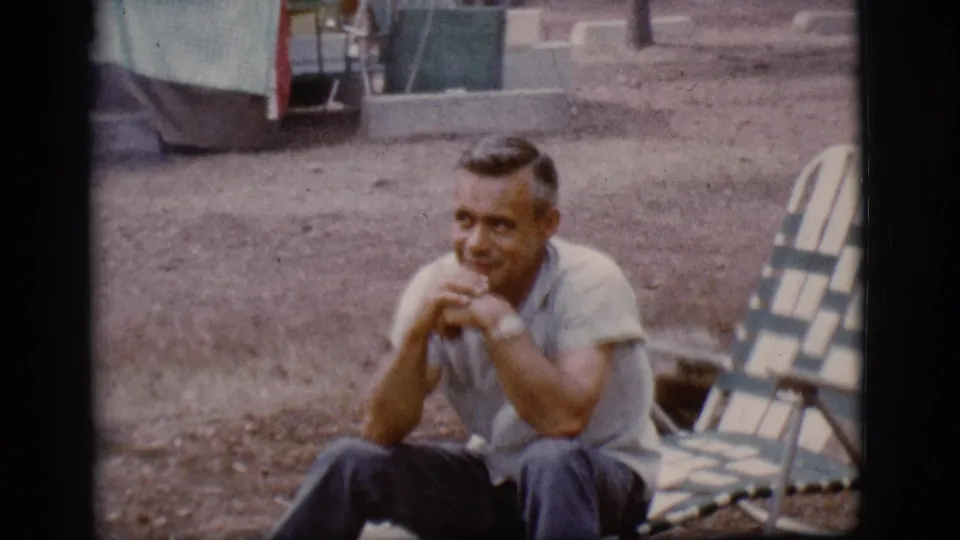 In supported 1968 Flagstaff home-movie footage, an adult man sits by a striped lounge chair near tent canvas on gravel ground.