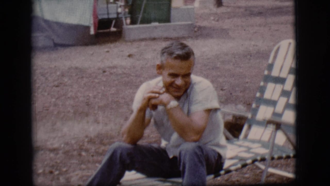 Seated man smiling beside a striped lounge chair on gravel.