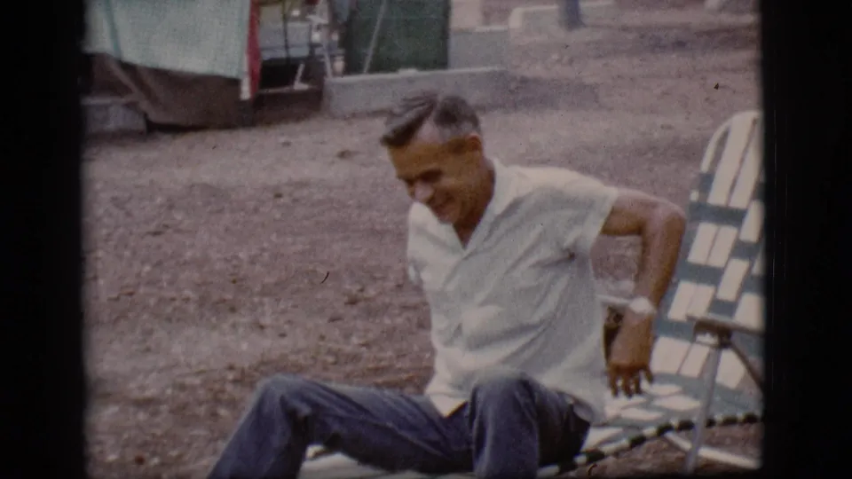 The man adjusts his seated position on gravel beside a striped lounge chair in the 1968 Flagstaff campsite scene.