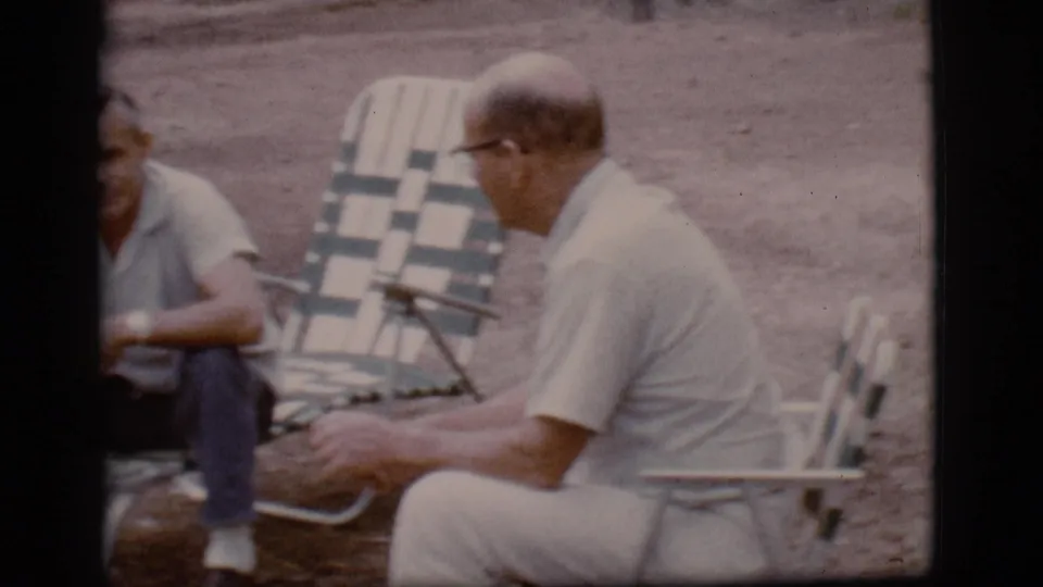 Two adult men sit near folding lounge chairs at a Flagstaff campsite, appearing to talk in candid 1968 home-movie footage.