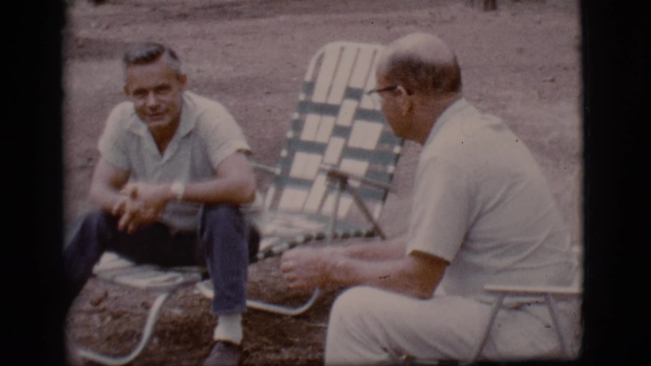 Two men sit outdoors in folding chairs on dirt ground, one facing the camera and one turned away.