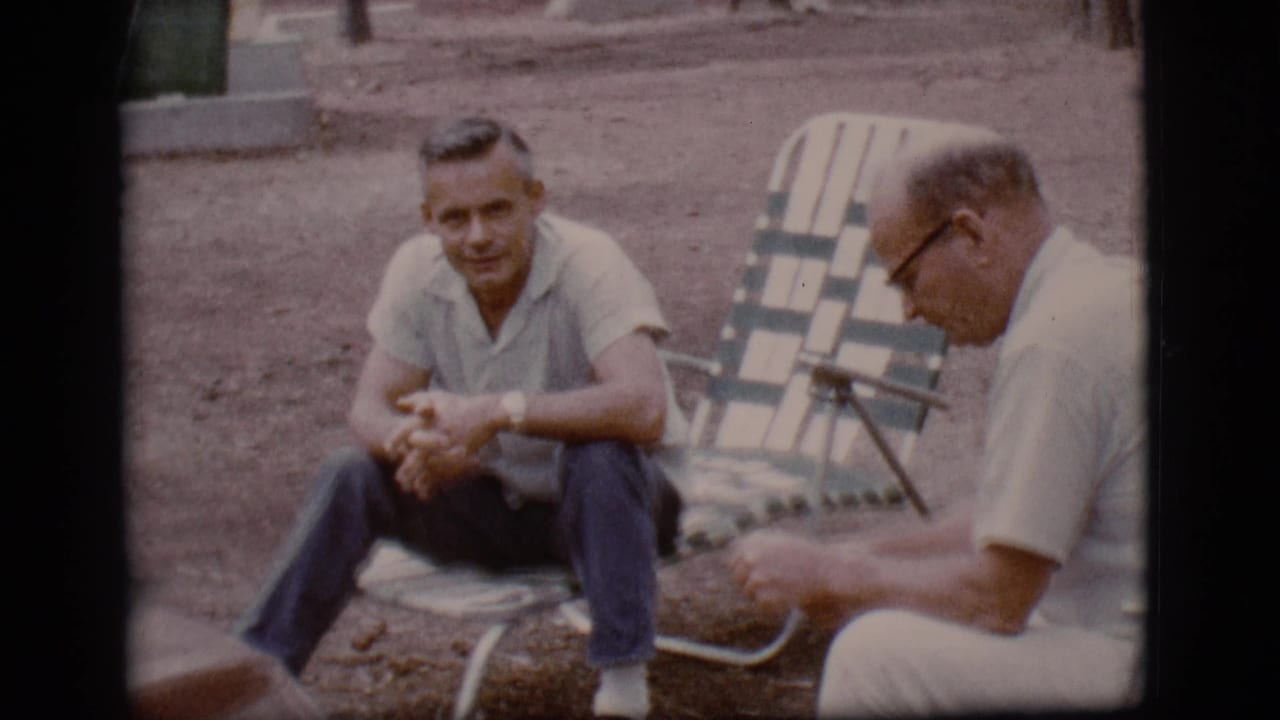 A man sits facing the camera while an older man in glasses sits beside him in a folding chair.