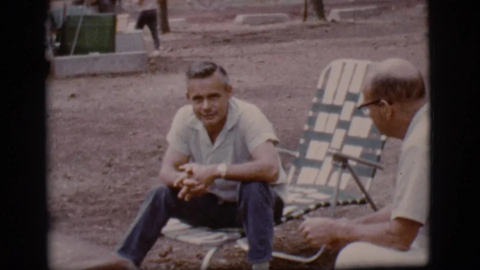 1968 Flagstaff frame of a man seated in a striped folding chair, with another man in glasses partly visible beside him.