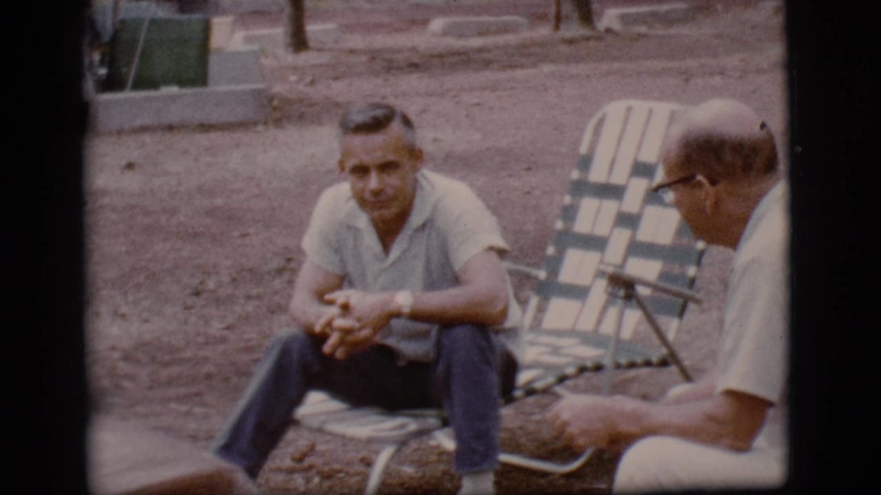 Two adult men sit on folding lawn chairs outdoors on dirt ground.