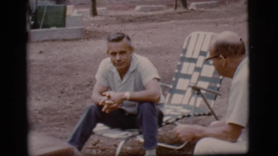 In 1968 Flagstaff, Arizona home-movie footage, two adult men sit facing each other on folding chairs at a campsite.