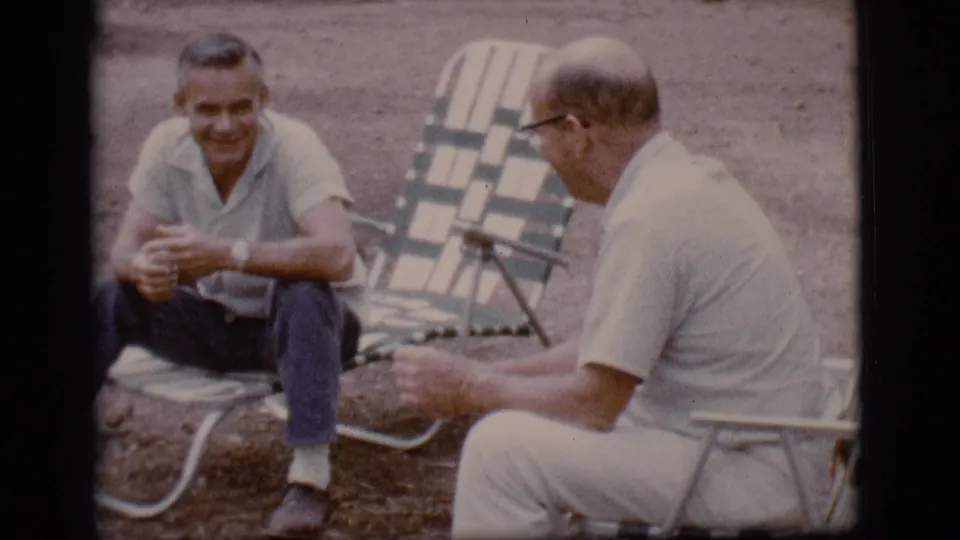 The 1968 home-movie frame shows one seated man smiling as he talks with another man in glasses at the campsite.