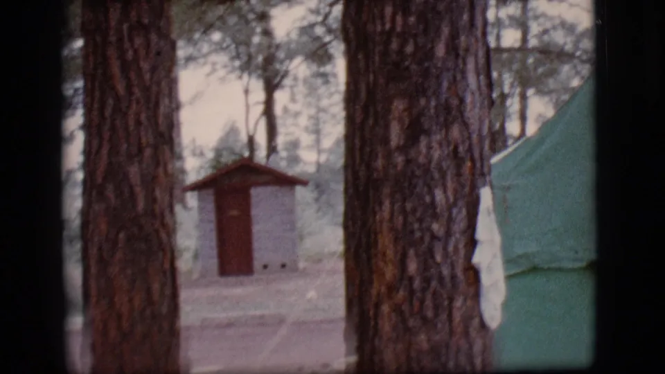 1968 Flagstaff home-movie frame showing pine trunks, a rustic campground outbuilding, and the edge of a green tent.