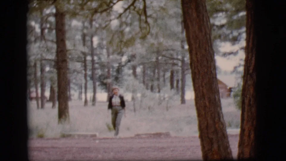 A person in casual outdoor clothing walks toward the camera through the 1968 wooded campground scene.