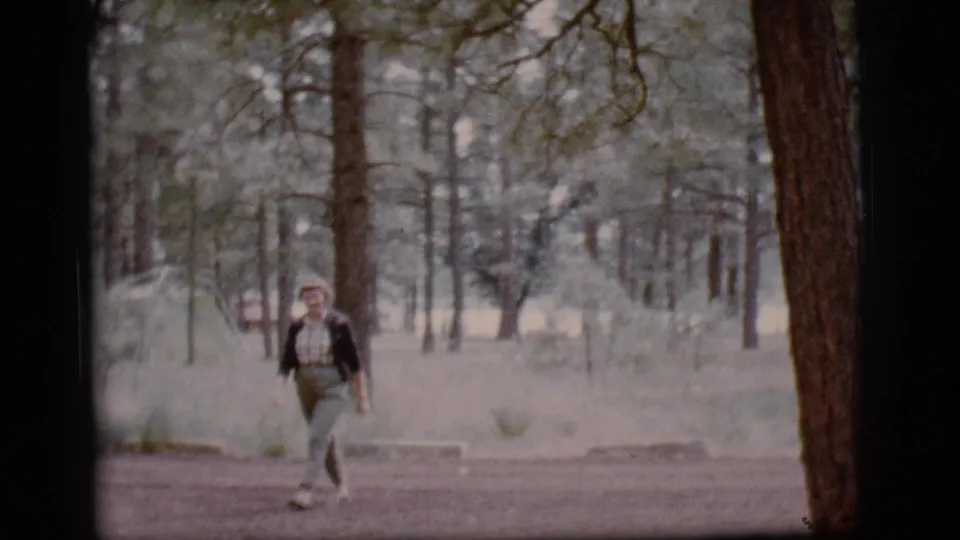 Smiling woman walks forward on a dirt path among pine trees in 1968 Flagstaff home-movie footage.
