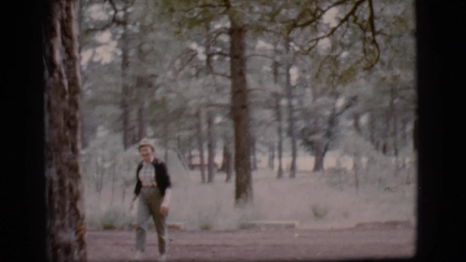 Young woman continues walking through a Flagstaff pine clearing, framed by tree trunks and soft 8mm color.