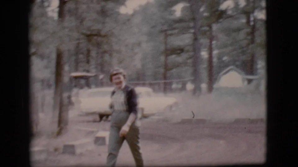 Woman walks past the camera in a Flagstaff forest setting, with a white car and cabin-like building in the background.