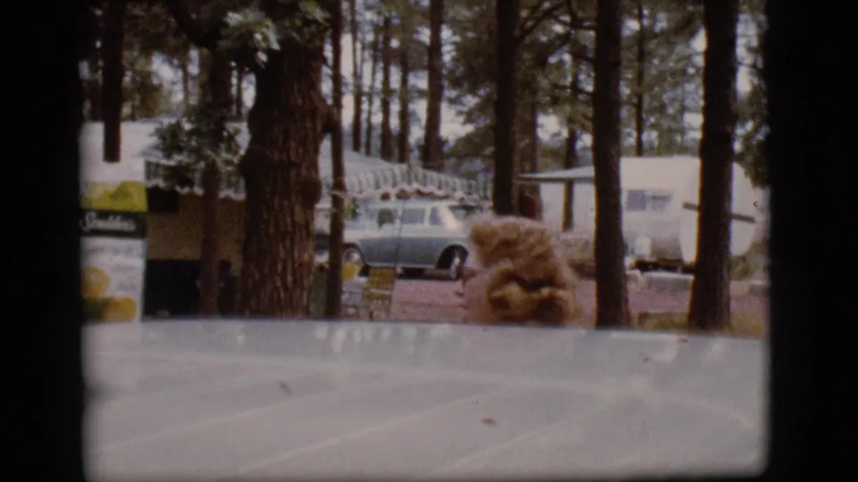The final frame looks across a vehicle surface toward pine trees, a parked vintage car, trailers, and a small brown object near the campsite.