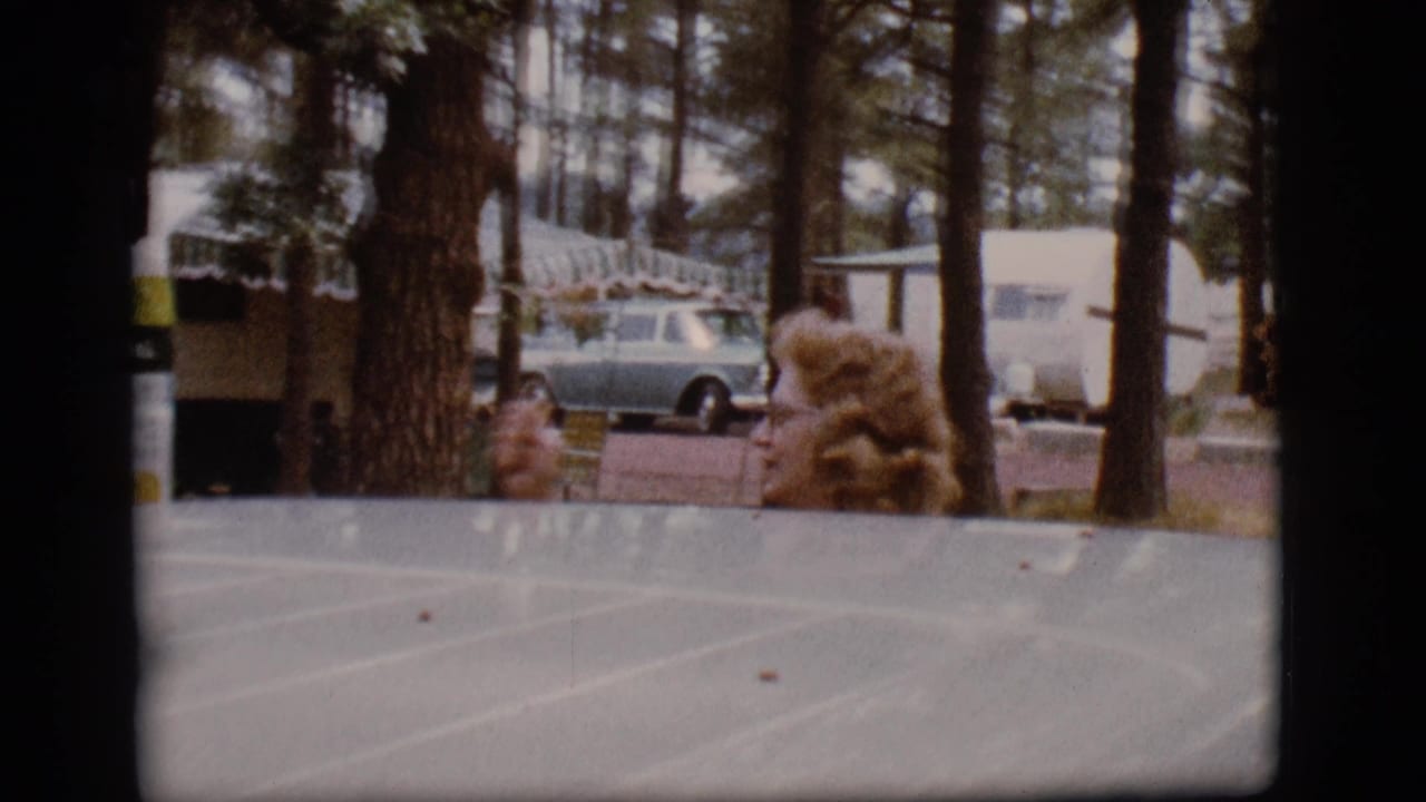 Woman with curly hair beside a vehicle roof at a wooded campground with trailers and a station wagon behind her.