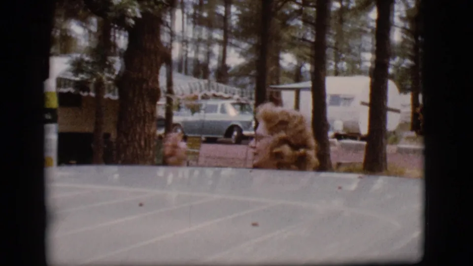 At a 1968 Flagstaff campground, a curly-haired woman stands partly hidden behind a vehicle roof with trailers and a light blue station wagon in the trees.