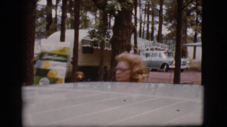 A woman wearing glasses is partly visible behind a vehicle roof in the Flagstaff campground, with trees, trailers, and a station wagon behind her.