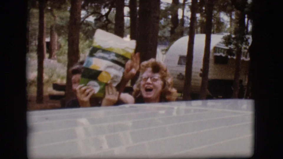 At the Flagstaff campground, the woman laughs and lifts a branded snack bag toward her face while another person stands nearby among trailers and trees.