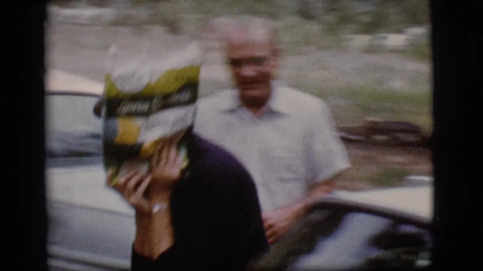 The woman covers her face with a branded snack bag at the 1968 Flagstaff campground as an older man stands near a car behind her.