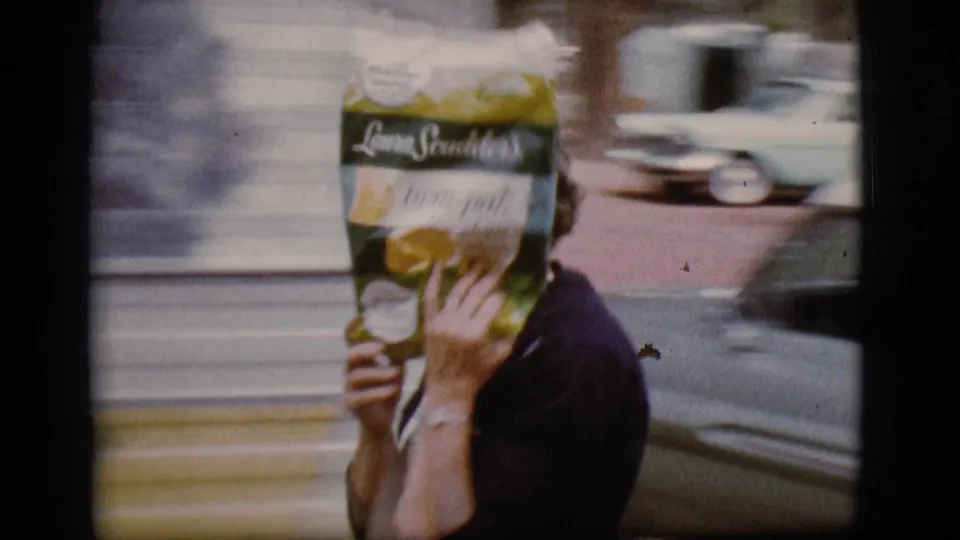 In supported 1968 Flagstaff home-movie footage, a woman in a dark top shields her face with a grocery bag near a camper trailer.