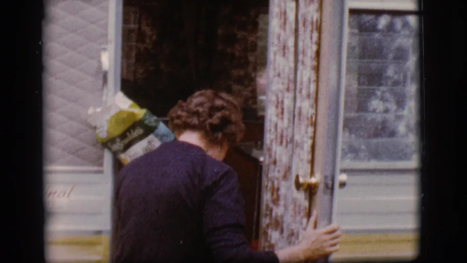 A woman with a grocery bag steps into a 1960s camper trailer doorway in Flagstaff, Arizona, as seen in color 8mm footage.