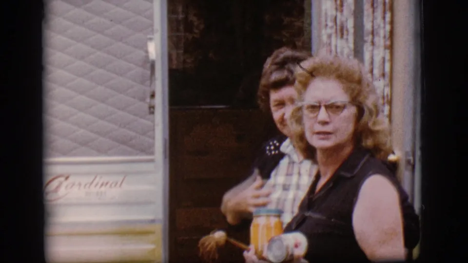 A curly-haired woman with glasses stands at the camper doorway holding a jar or container, with another person partly visible behind her.
