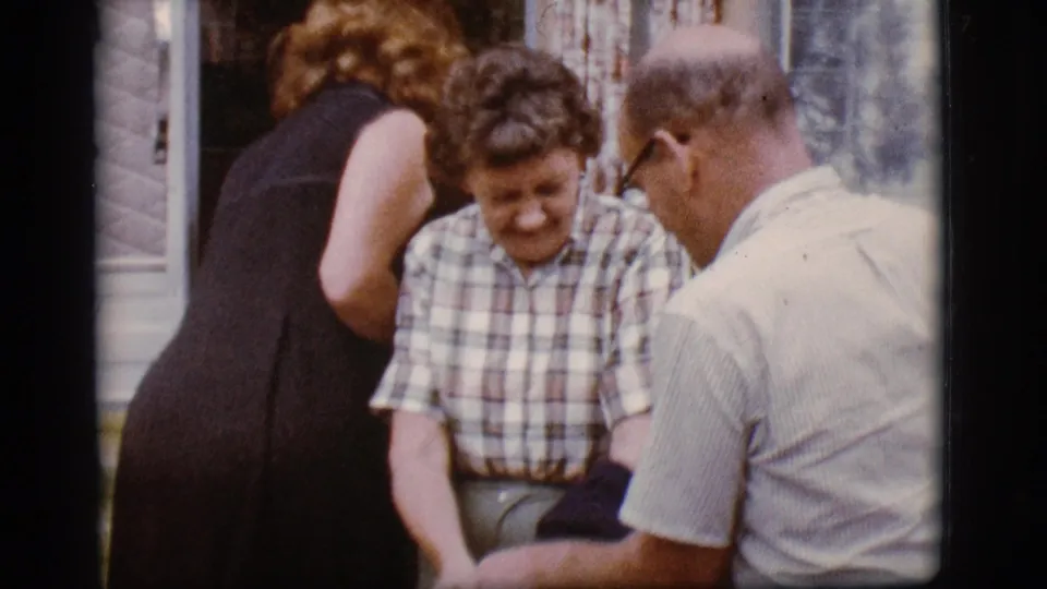 Three adults gather outside a mobile home in the 1968 Flagstaff home movie, with the woman in plaid smiling while a man in a light shirt faces her.