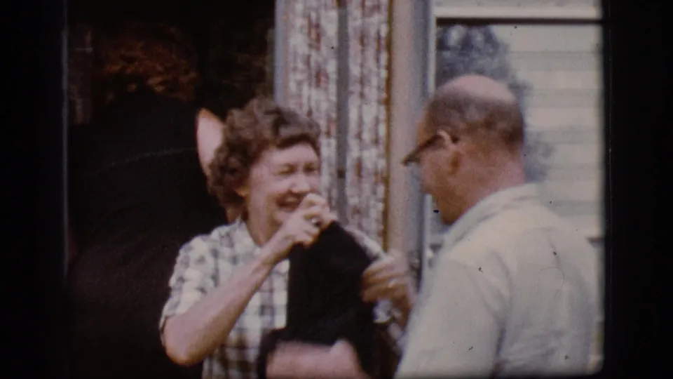 A woman in plaid laughs and holds a dark garment near a man in a light shirt outside the mobile home in this 1968 Flagstaff home-movie frame.