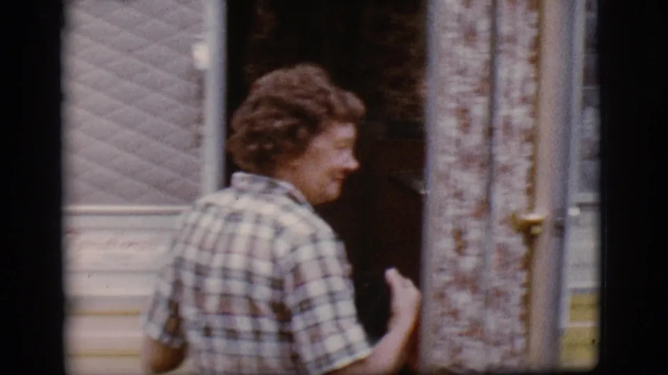The woman in a plaid shirt turns into the camper doorway, with the yellow-and-white exterior and window visible around her.